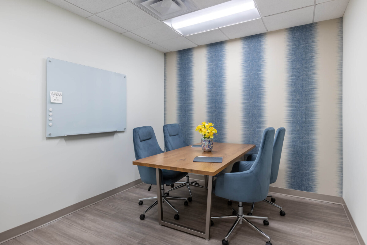 ?seraph accel gi=wp content%2Fuploads%2F2024%2F07%2F008 FD 1 - Metro Offices Small meeting room with a wooden table, four blue upholstered chairs, a whiteboard on the left wall, and blue striped accent wallpaper on the right. A vase with yellow flowers is on the table.