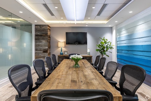 ?seraph accel gi=wp content%2Fuploads%2F2024%2F07%2F010 RR - Metro Offices A modern conference room with a long wooden table, eight black mesh chairs, a wall-mounted screen, a plant, and a lamp. The walls feature glass panels and blue accents.