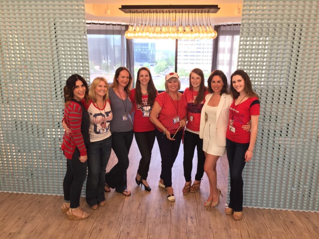 KB Broker Event Greensboro Station - Metro Offices A group of eight women smiling and posing together in the Metro Offices with a city view, several wearing red and white clothing likely supporting a sports team or event.