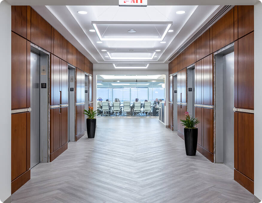 Modern office hallway with four elevators, wood paneling, two potted plants, and a view into a glass-walled conference room with people seated inside.