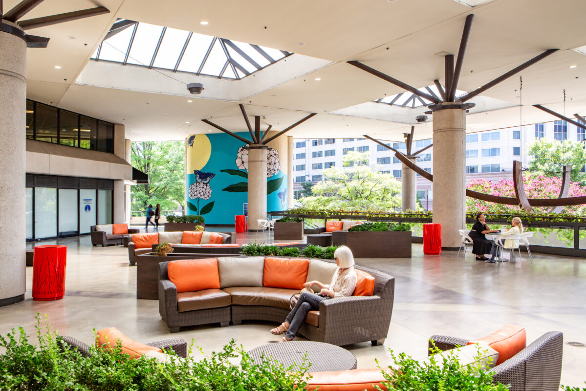 The outdoor seating area at the office space in Chevy Chase features modern furniture with orange cushions. Visitors relax or stroll among plants and pillars, beneath skylights that flood the vibrant mural-covered walls with natural light.