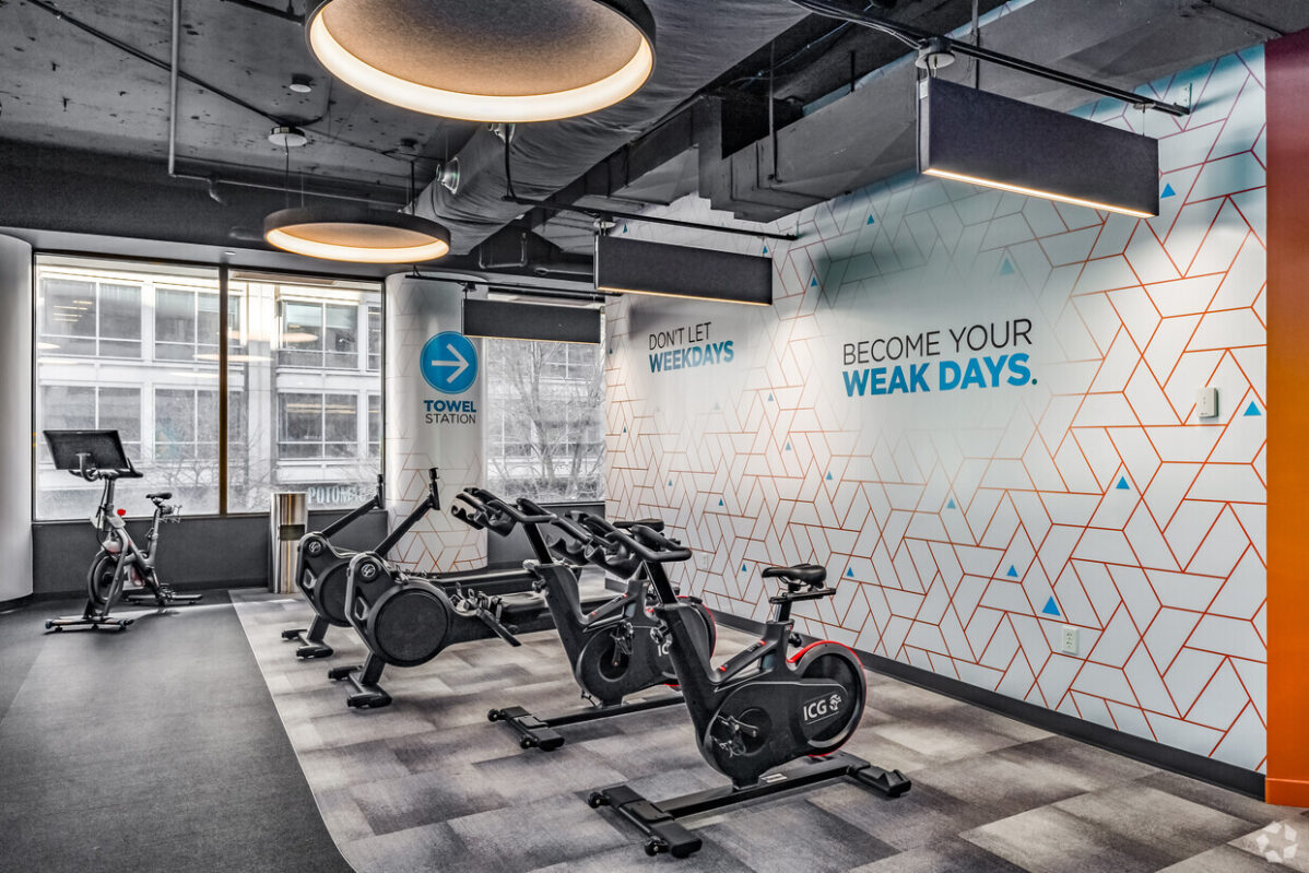 The gym room, nestled within an office space in Chevy Chase, features three stationary bikes and a wall adorned with motivational text. A towel station sign adds convenience while large windows welcome natural light. Modern ceiling lights ensure the space is brilliantly illuminated.