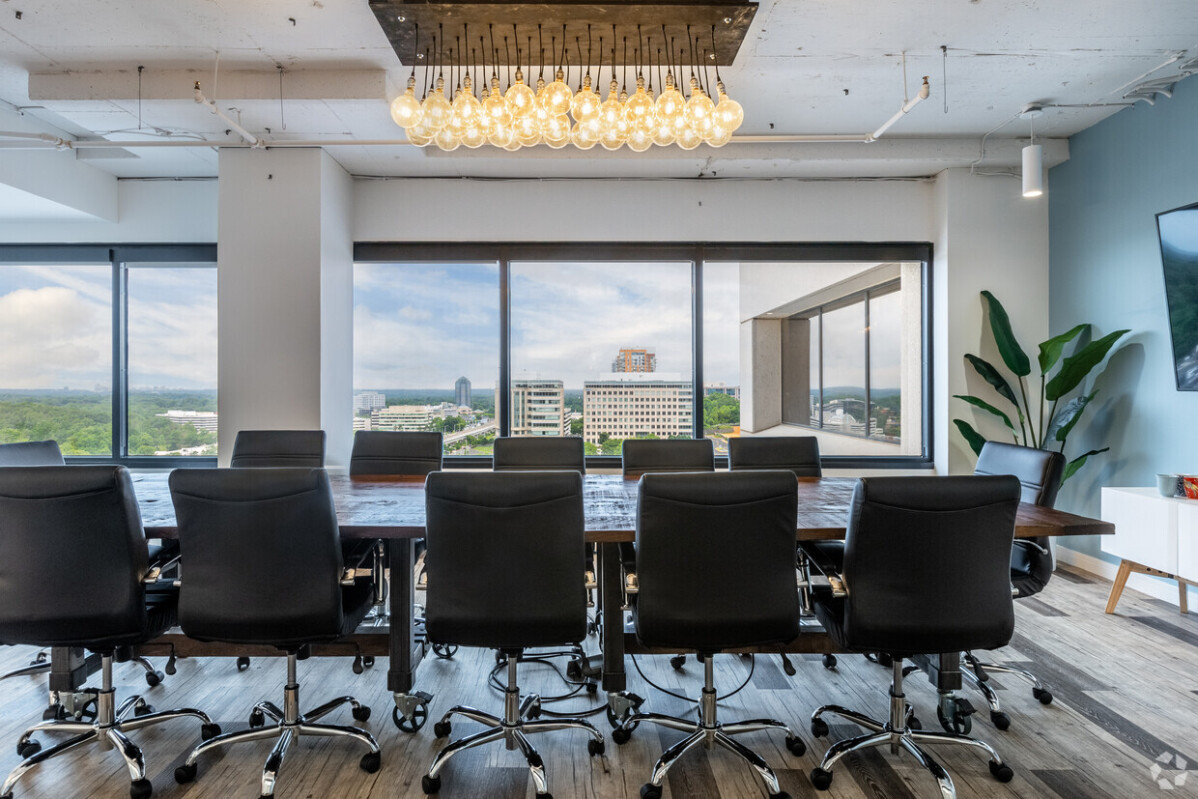 A modern conference room featuring the Vista wooden table, surrounded by black leather chairs, a large window offering a stunning city vista, and an elegant pendant light fixture overhead.