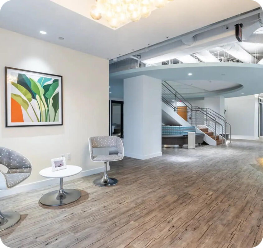 Modern office lobby with two gray chairs, a small round table, abstract plant artwork on the wall, wood flooring, and a staircase leading to an upper level—an inviting space to meet or make contact in style.