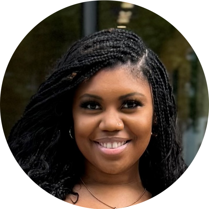 A woman with braided hair smiles at the camera, standing outdoors with a blurred background.