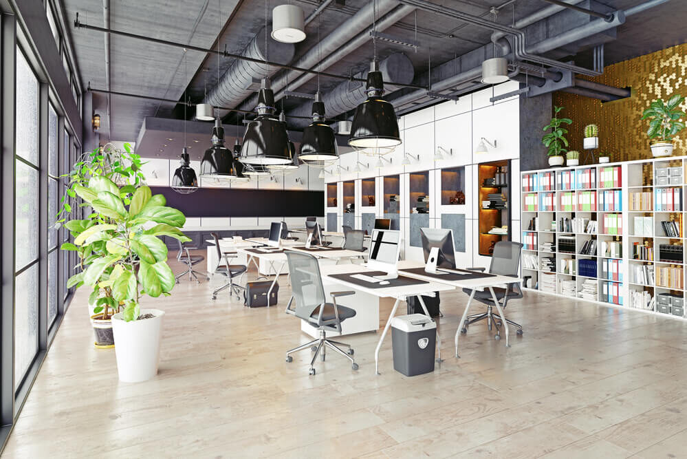 Modern open-plan office designed for collaboration, featuring multiple desks, computers, ergonomic chairs, large windows, shelves with files, potted plants, and exposed ceiling ductwork—ideal for vibrant coworking spaces.