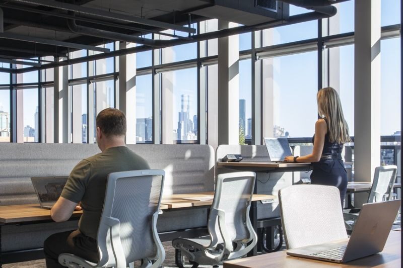 Two people work at desks in a modern office with large windows and a city skyline view, embracing hybrid work and utilizing AI productivity tools to enhance their workflow in 2026.