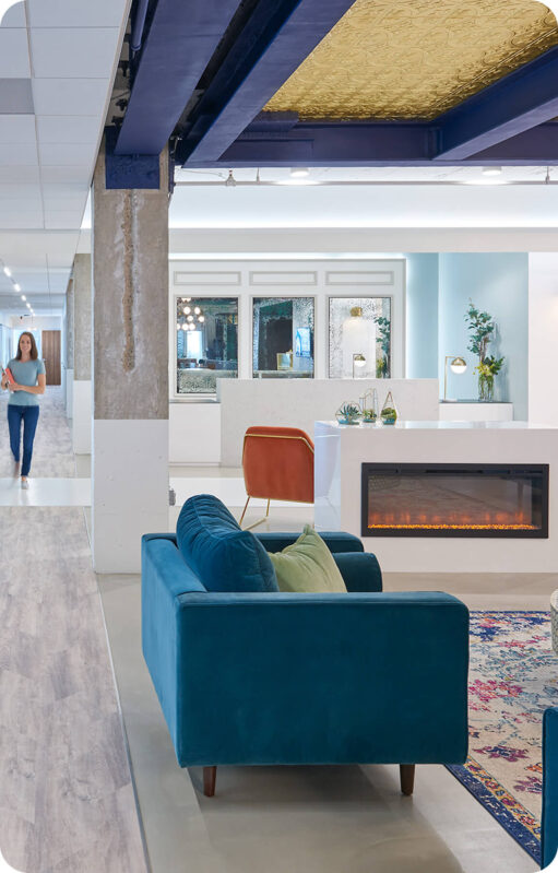 A modern office lounge with a blue sofa, orange chair, electric fireplace, and a woman walking down a hallway in the background—perfect for relaxed meetings or casual contact with colleagues.
