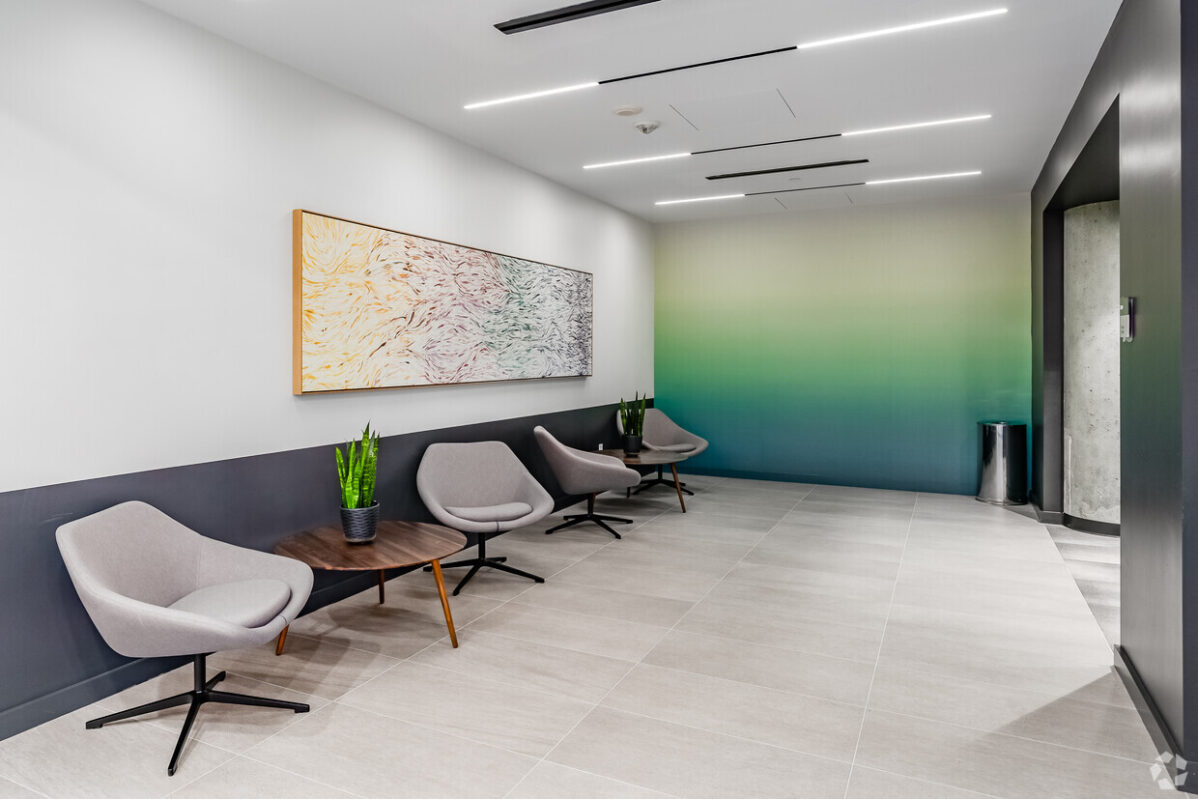 Modern lobby in an office space in Chevy Chase, featuring gray chairs, small tables, and potted plants. Abstract art adorns a wall with a gradient color design. The minimalistic and well-lit area is complemented by sleek tiled flooring.