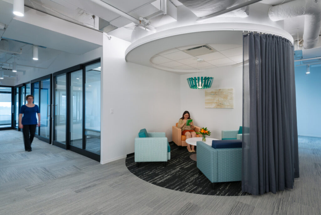 A modern office lounge area with three teal armchairs around a white table, partially enclosed by a circular curtain. One person is sitting and reading, evaluating some documents, while another walks in the hallway, reflecting the dynamic nature of shared environments often found in coworking spaces.