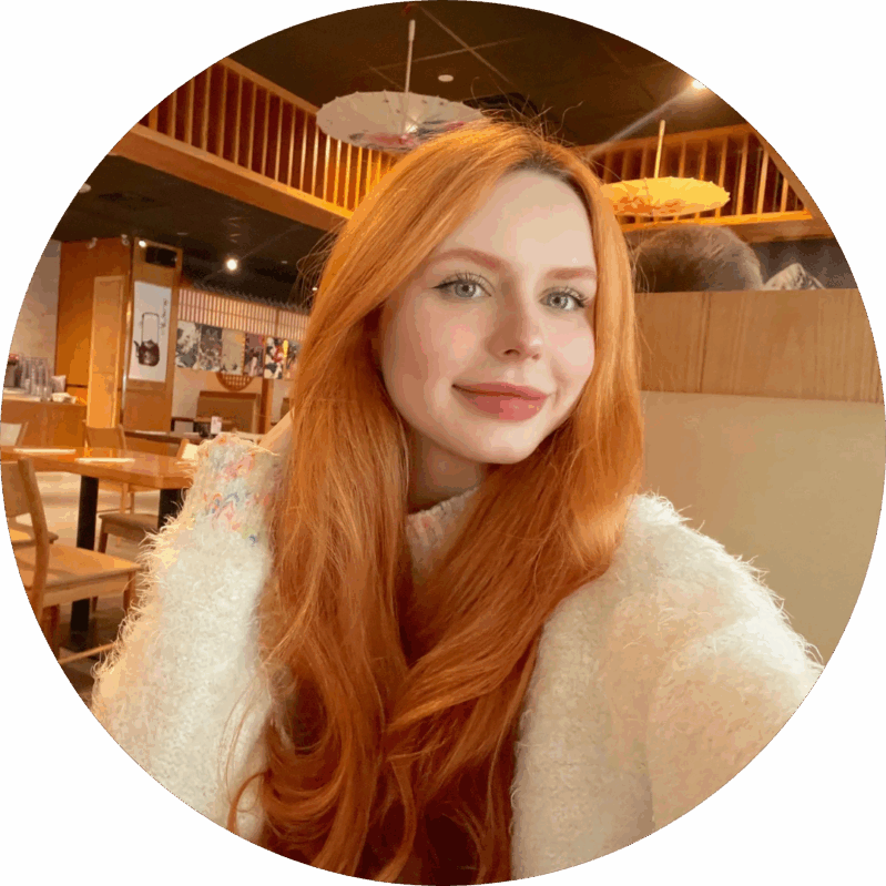 A woman with long red hair and a white sweater takes a selfie in a restaurant with wooden decor and parasols on the ceiling.