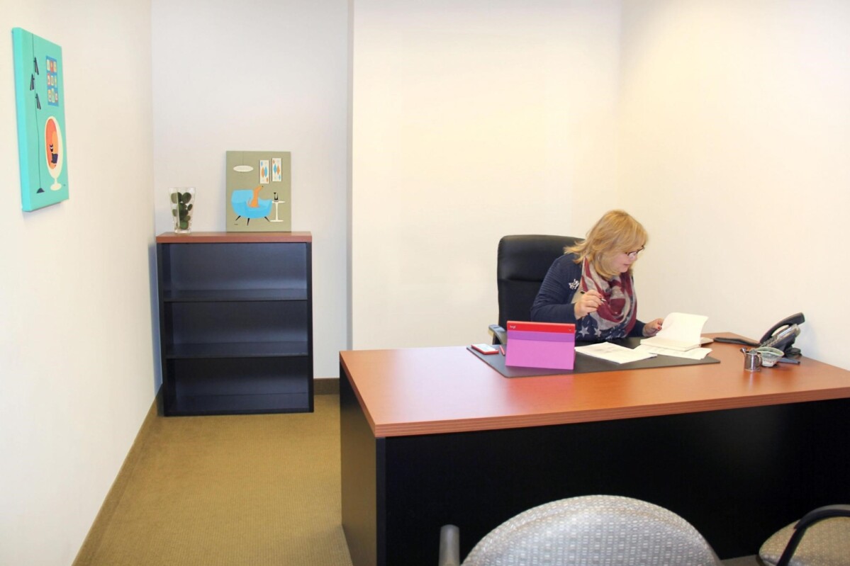 ?seraph accel gi=wp content%2Fuploads%2F2024%2F07%2F004 DO - Metro Offices A woman sits at a desk in an office with a phone, papers, a laptop, and a bookshelf. The walls are adorned with two pieces of colorful art.
