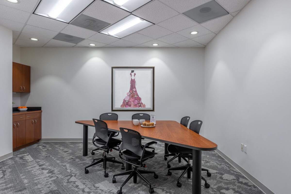 ?seraph accel gi=wp content%2Fuploads%2F2024%2F07%2F004 MC - Metro Offices A conference room with a long wooden table, five black swivel chairs, a framed painting on the wall, and cabinets in the corner. Bowl of fruit and informational stand on the table.