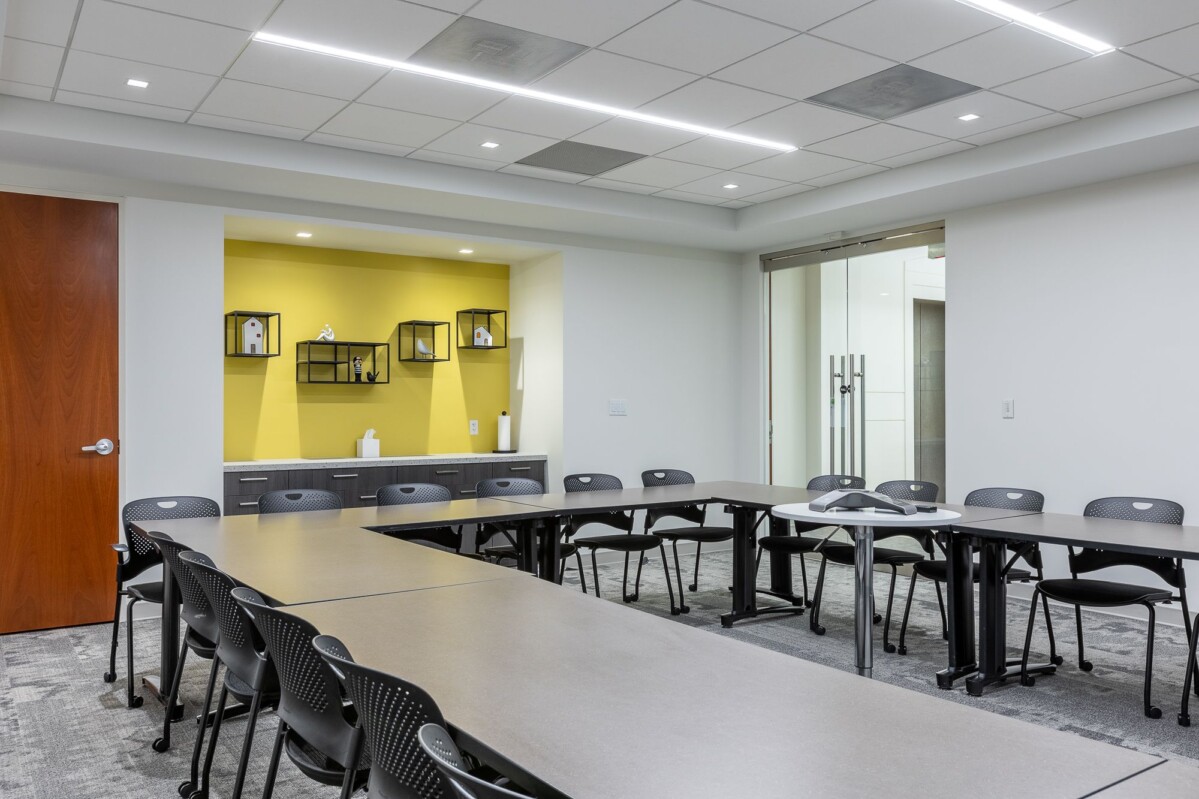 ?seraph accel gi=wp content%2Fuploads%2F2024%2F07%2F004 TR - Metro Offices The Shenandoah Training Room boasts a contemporary conference environment, complete with U-shaped rectangular tables, numerous black chairs, a wooden door, and a yellow accent wall adorned with shelves.