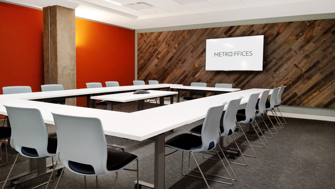 ?seraph accel gi=wp content%2Fuploads%2F2024%2F07%2F005 FD - Metro Offices A modern conference room with a large U-shaped table, several gray chairs, a TV on the wall displaying "Metro Offices," and orange and wooden decor.