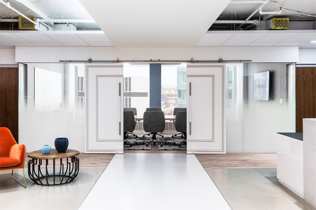 ?seraph accel gi=wp content%2Fuploads%2F2024%2F07%2F005 LC - Metro Offices Modern office space with a corridor leading to a glass-walled conference room furnished with gray chairs. The corridor has a white ceiling and floor, with an orange chair and side table on the left side.