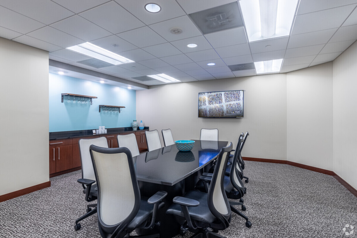 ?seraph accel gi=wp content%2Fuploads%2F2024%2F07%2F006 MC - Metro Offices A modern conference room with a long black table, eight white office chairs, a wall-mounted TV, shelves, and a blue decorative bowl on the table. The room has overhead lighting and carpeted flooring.
