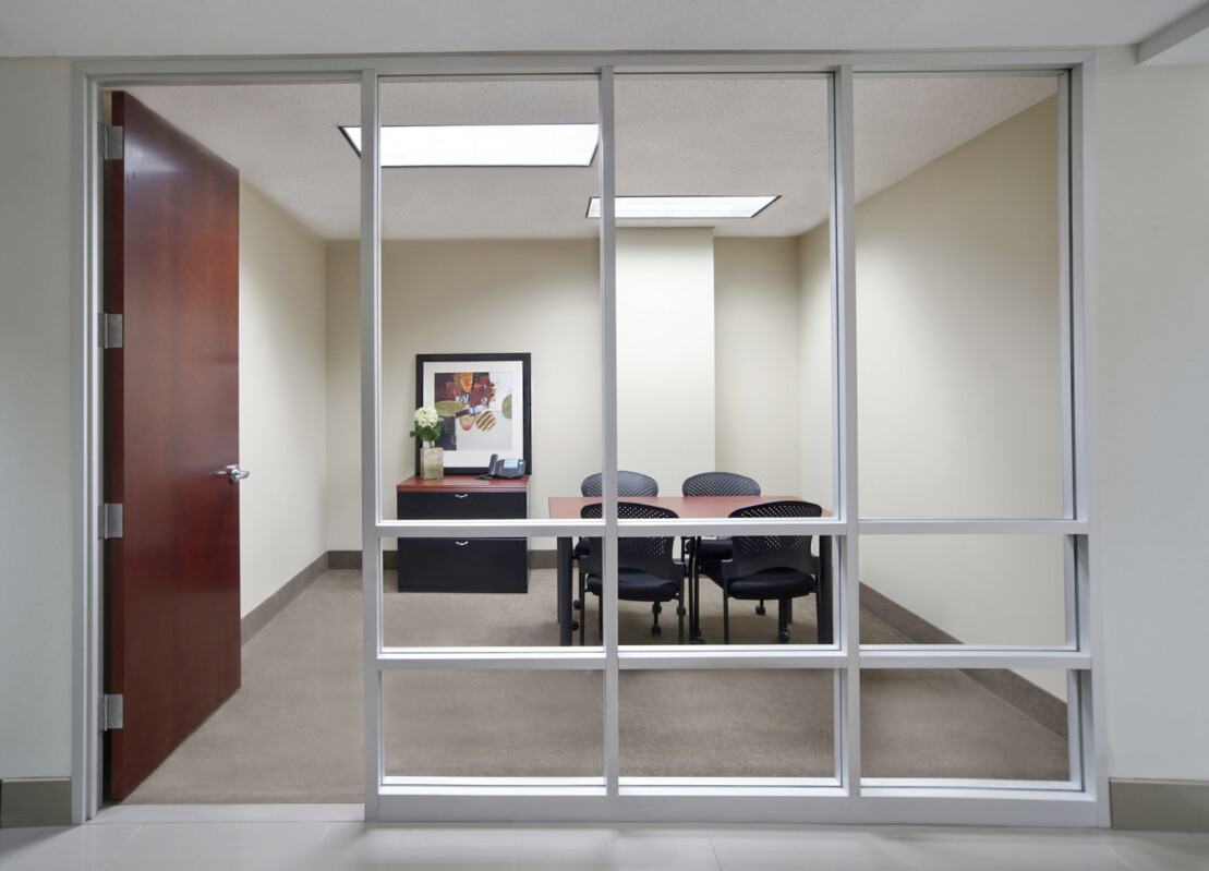 ?seraph accel gi=wp content%2Fuploads%2F2024%2F07%2F006 SC - Metro Offices A modern office meeting room with a glass wall, a wooden door, a framed picture on the wall, a desk with flowers and a phone, and five chairs around a conference table.