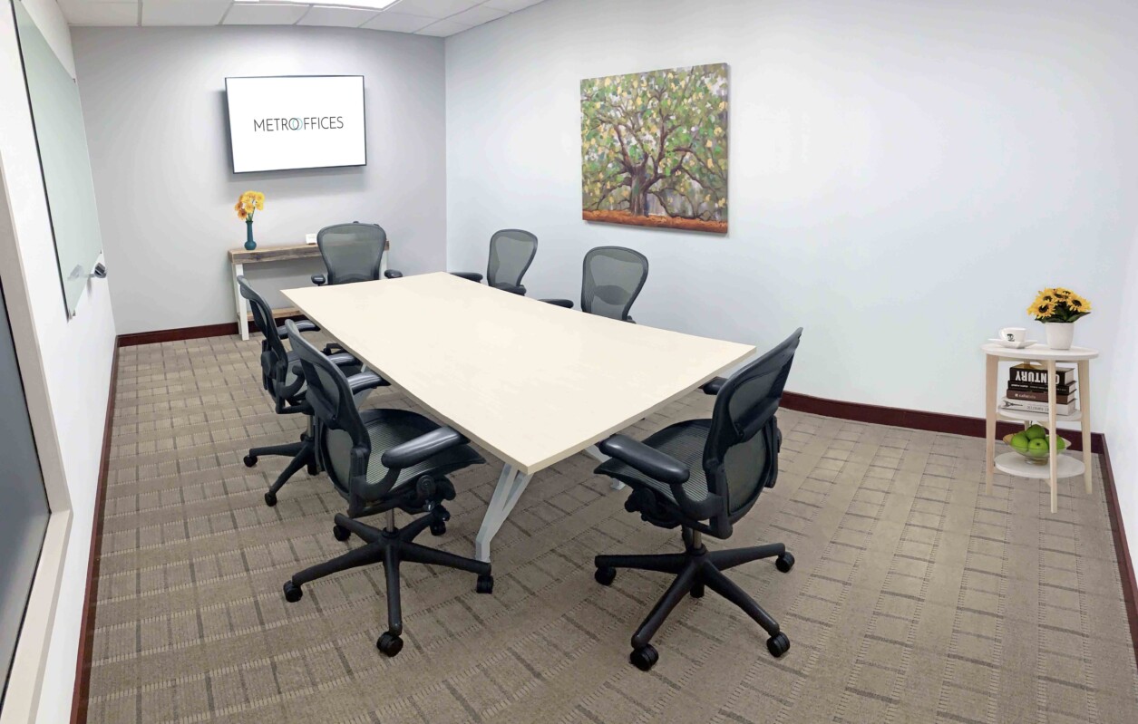 ?seraph accel gi=wp content%2Fuploads%2F2024%2F07%2F007 MC - Metro Offices A small conference room with a rectangular table, six office chairs, a wall-mounted screen showing "Metro Offices," a painting, and a side table with sunflowers and a green fruit basket.