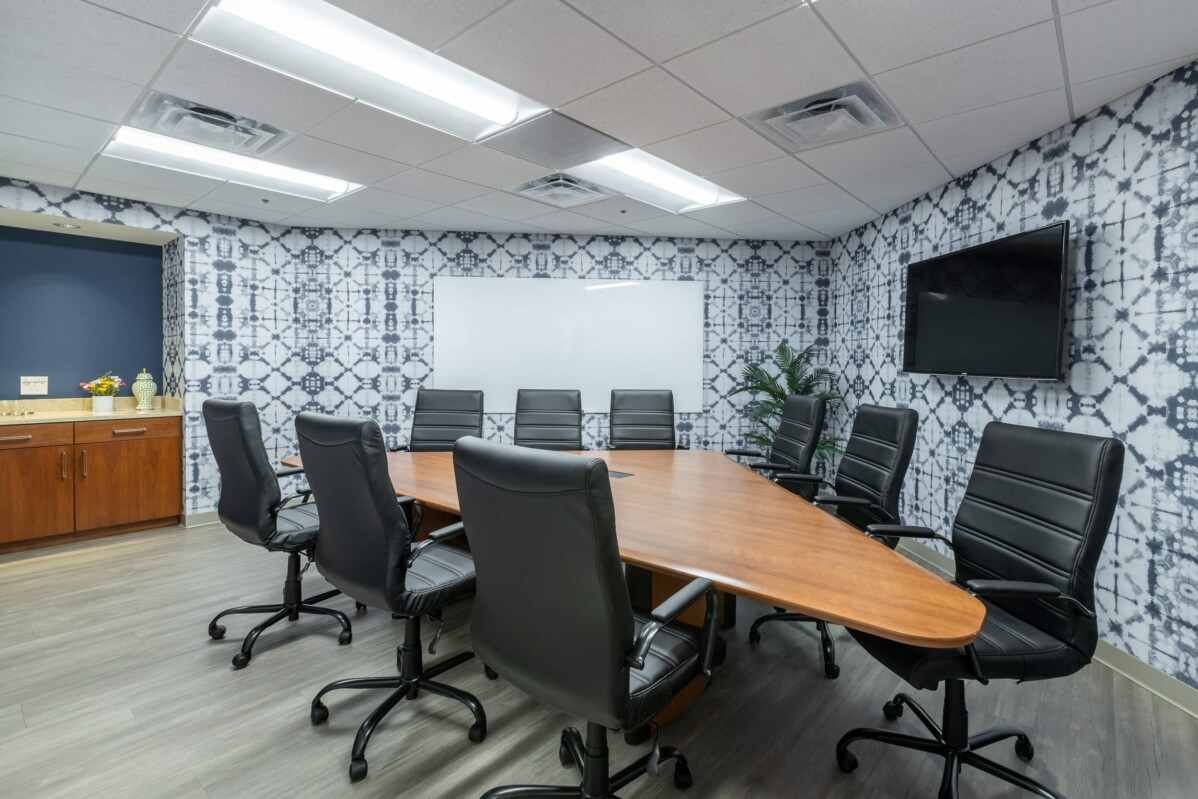 ?seraph accel gi=wp content%2Fuploads%2F2024%2F07%2F008 LC2 - Metro Offices A conference room with a large oval wooden table surrounded by eight black office chairs, a TV mounted on the wall, and a whiteboard. The walls are patterned and a cabinet is in the corner.