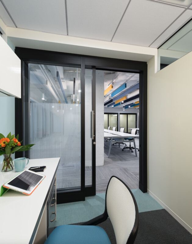 ?seraph accel gi=wp content%2Fuploads%2F2024%2F07%2F009 DO - Metro Offices An office cubicle with a desk, chair, tablet, and flowers. A large glass sliding door separates the cubicle from a conference room with chairs and a long table.