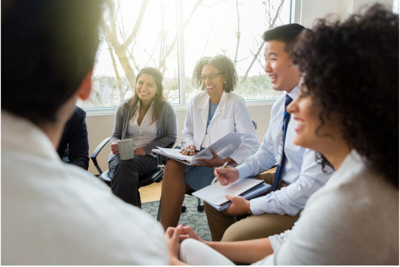 Healthcare - Metro Offices A group of professionals sits in a circle, holding notepads and smiling, engaged in a discussion in a brightly lit room.
