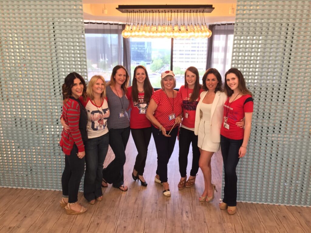 A group of eight women smiling and posing together in the Metro Offices with a city view, several wearing red and white clothing likely supporting a sports team or event.